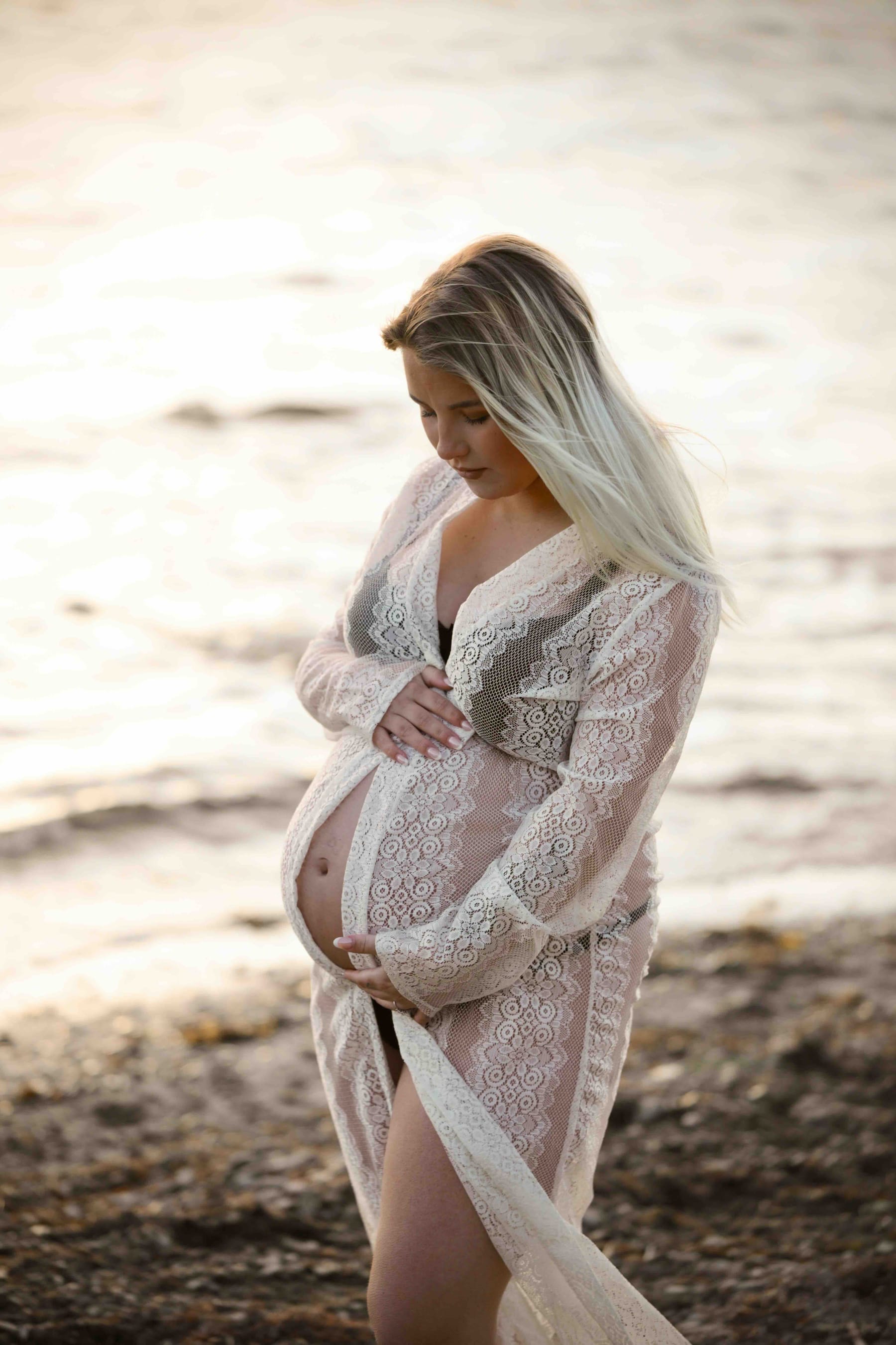Gravid kvinna vid havet i spetsklänning i solnedgång